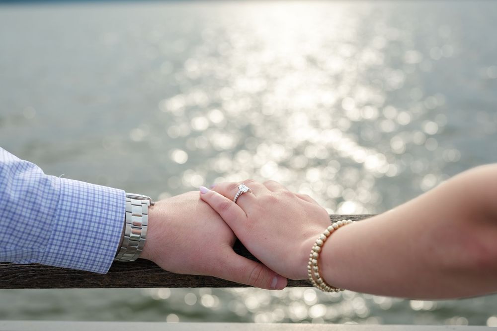 Couple holding hands on a wooden railing by the water, woman wearing a pearl bracelet and engagement ring.