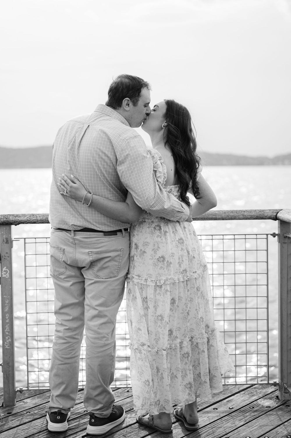 Black and white photo of a couple sharing a romantic kiss on a waterfront pier, the woman in a floral dress.