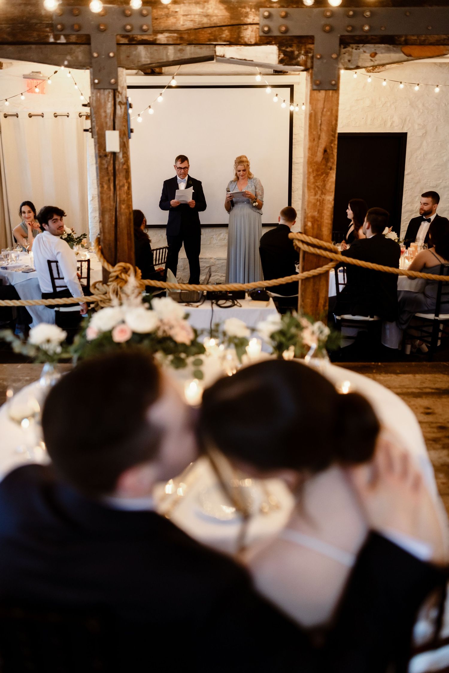 Wedding reception speech in a rustic barn venue with string lights and exposed wooden beams.
