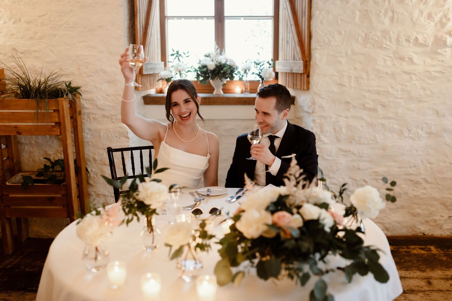 Newlyweds raise glasses in celebration at their reception table decorated with white floral arrangements.