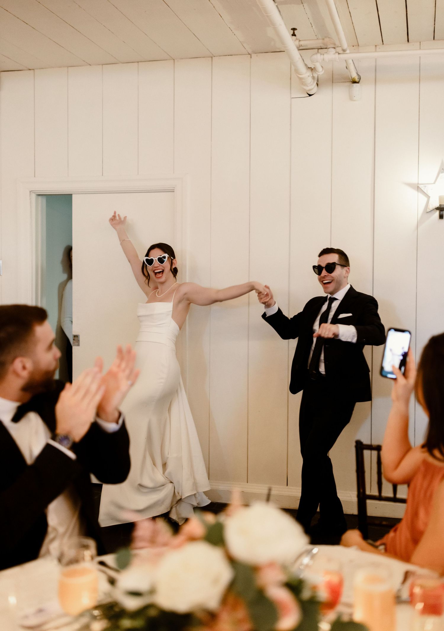 Wedding couple wearing sunglasses dance energetically at their reception in white rustic venue.