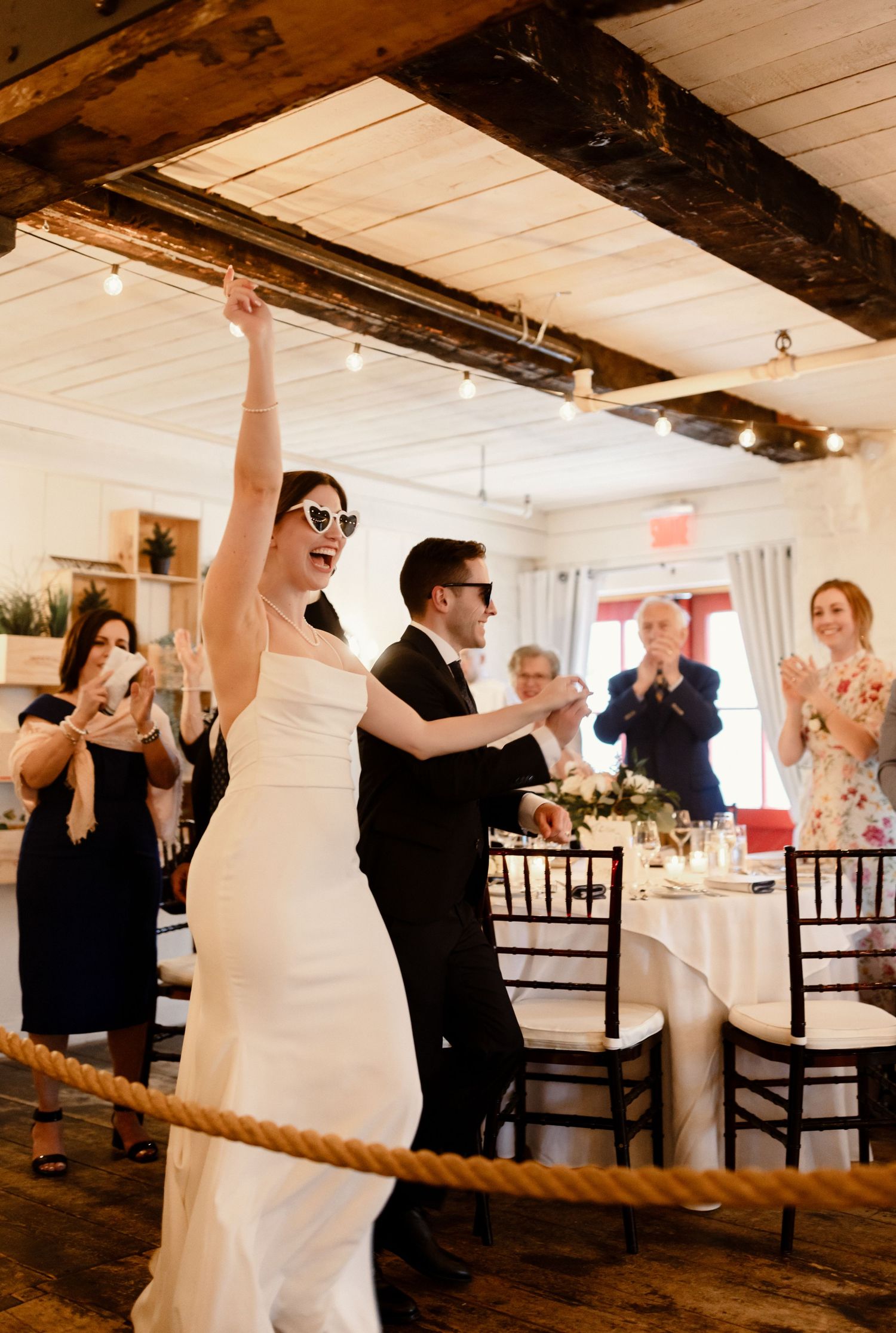 Wedding celebration in rustic white venue with guests cheering and string lights overhead.