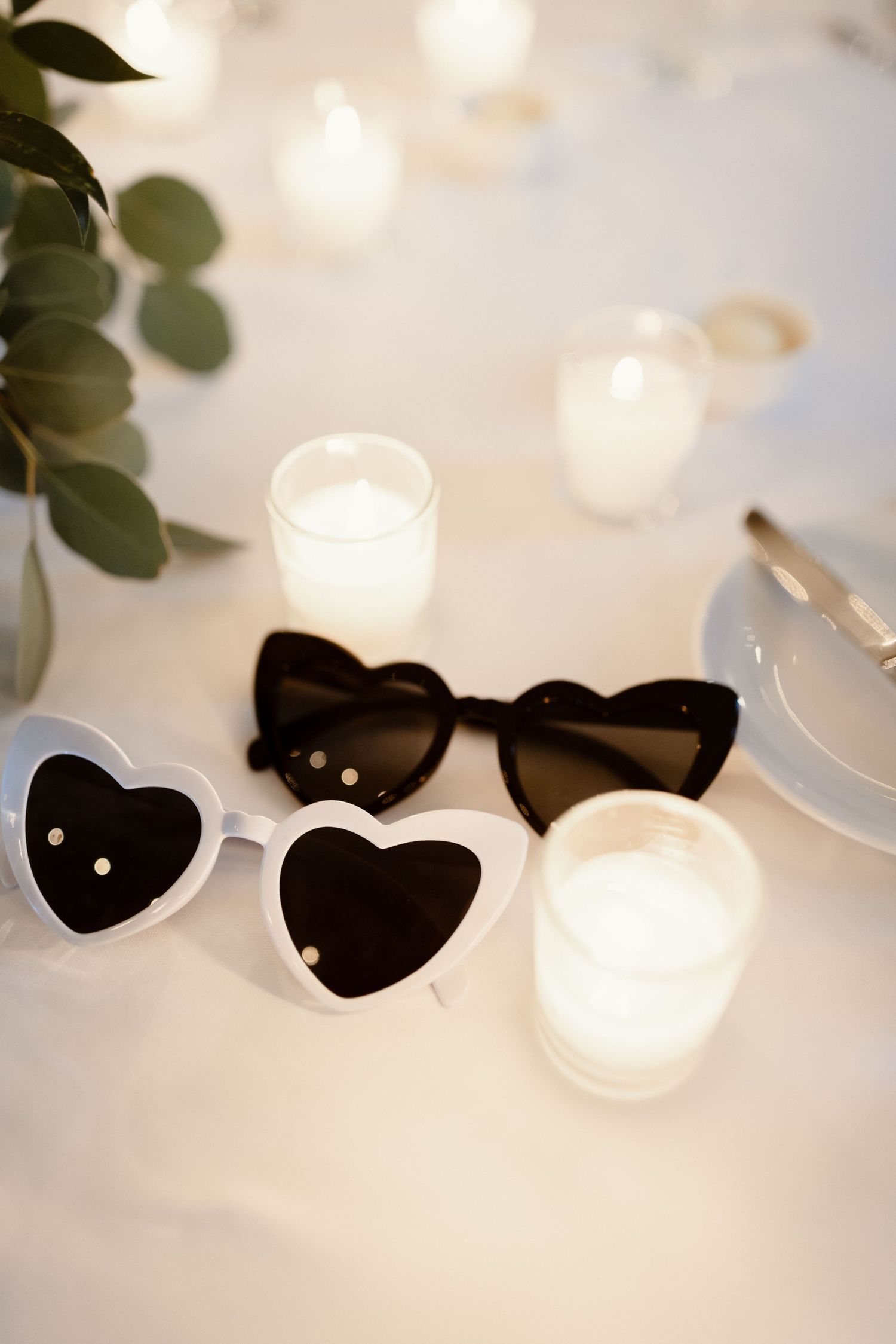 Heart-shaped sunglasses and tea light candles arranged on white tablecloth at wedding reception.