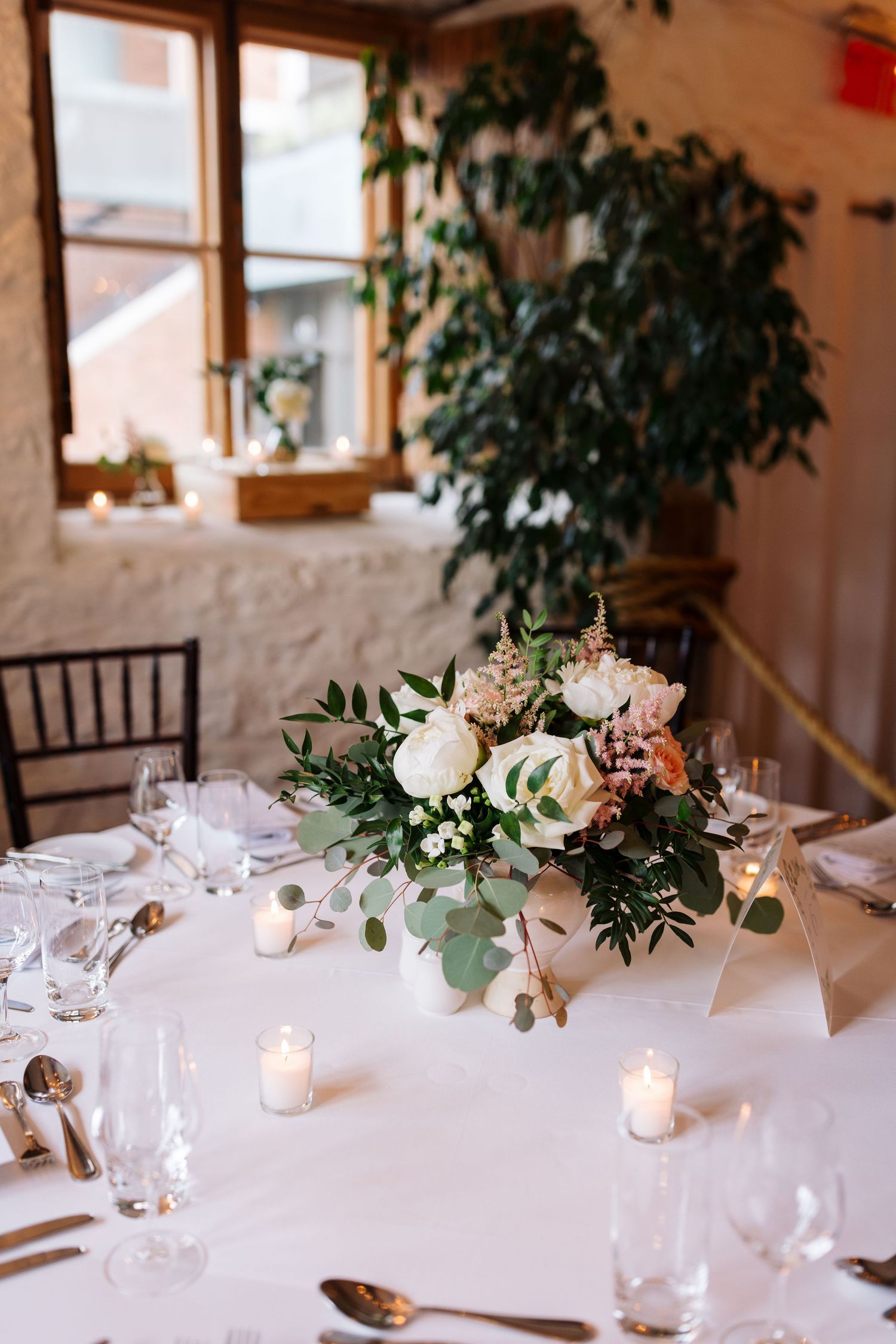 White and peach floral centerpiece with greenery on an elegantly set table near a bright window.
