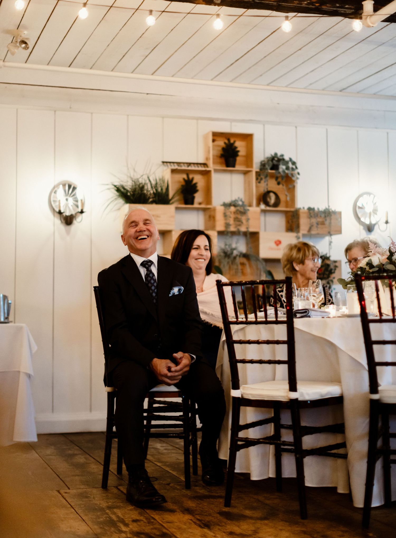 Wedding guests smile and laugh while seated at elegantly decorated reception tables with white linens and rustic decor.