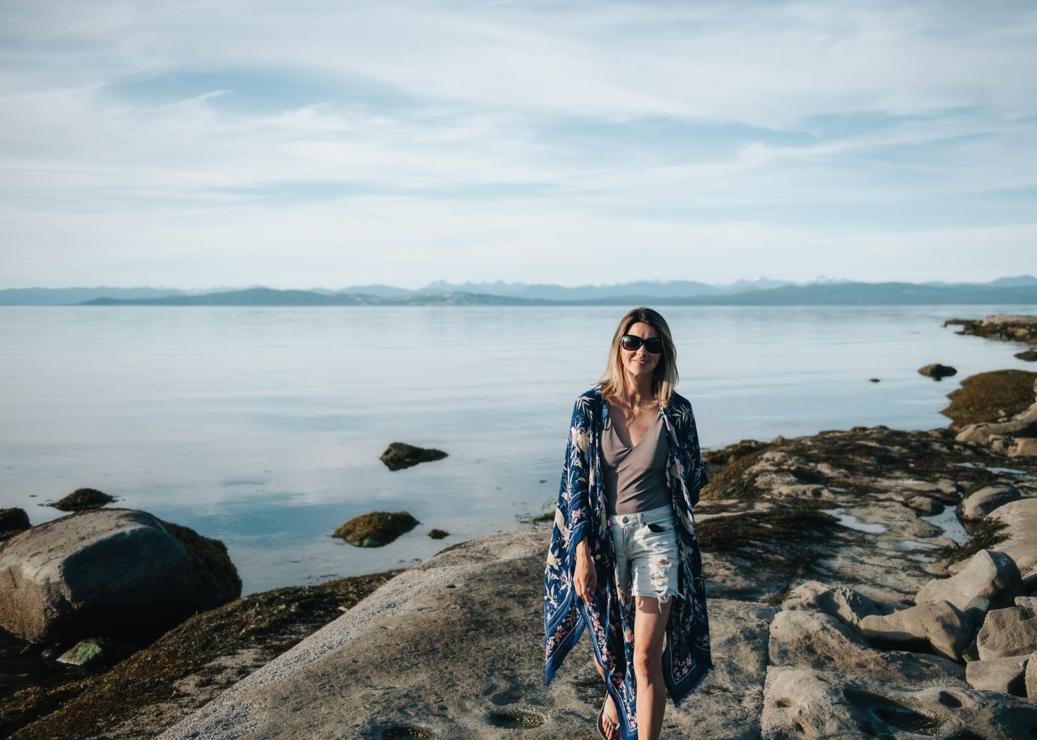 Person in flowy dress walks along rocky coastal shoreline with mountains in background.