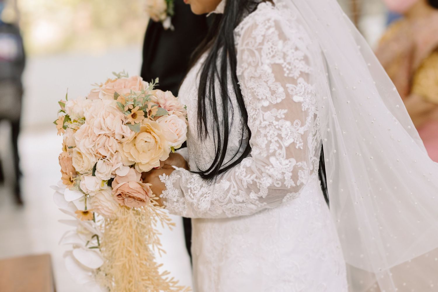 Close-up of a bride in lace sleeves holding a blush-toned cascading bouquet.