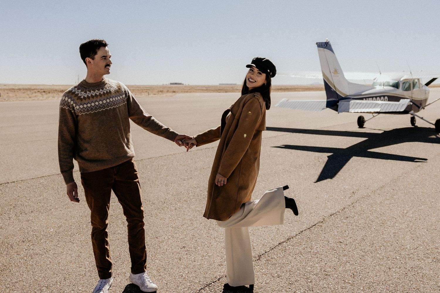 Couple holding hands on airport tarmac with small private plane in background on sunny day.