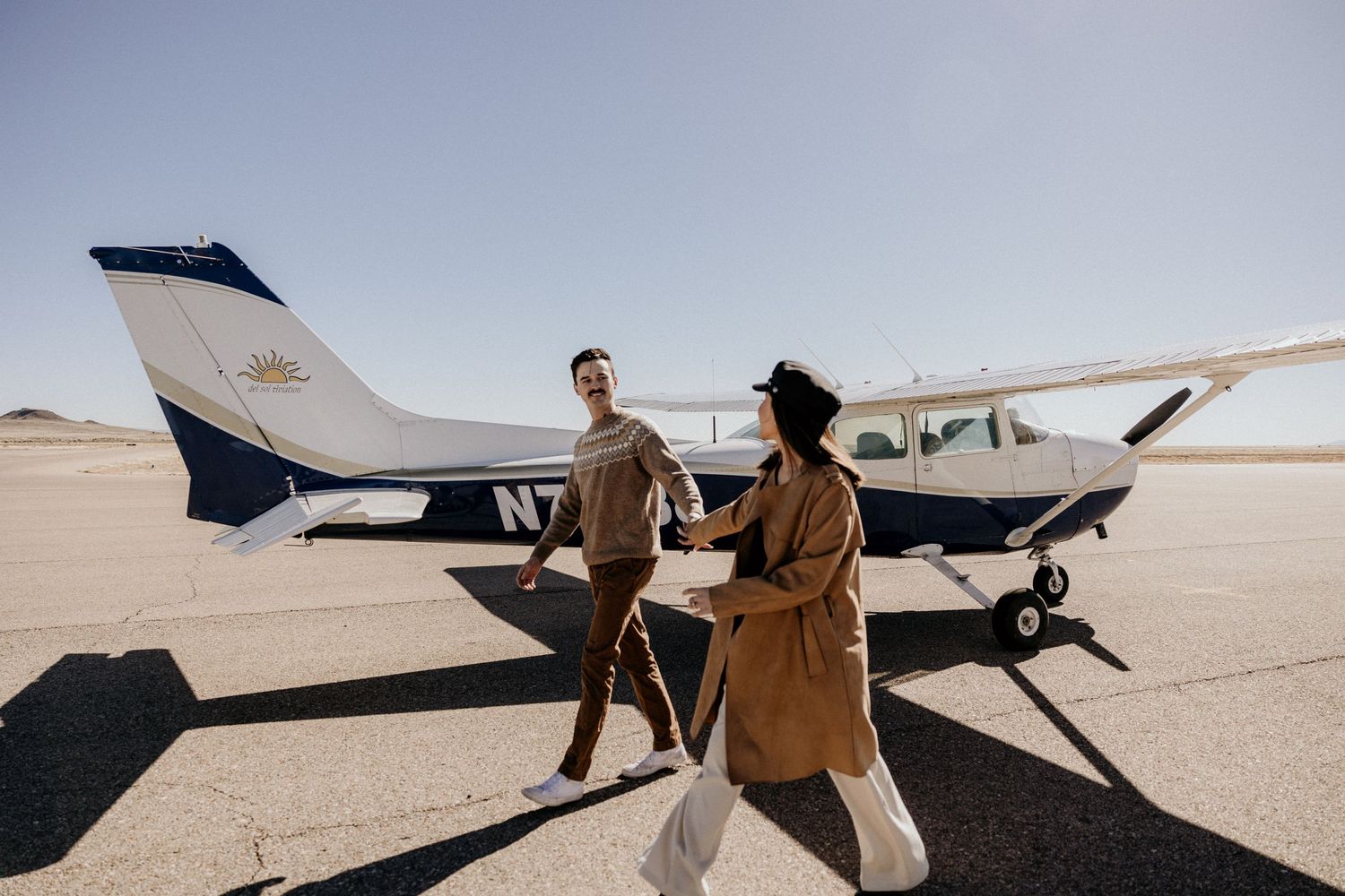 A couple walks hand-in-hand past a small private Cessna airplane on a sunny airstrip.