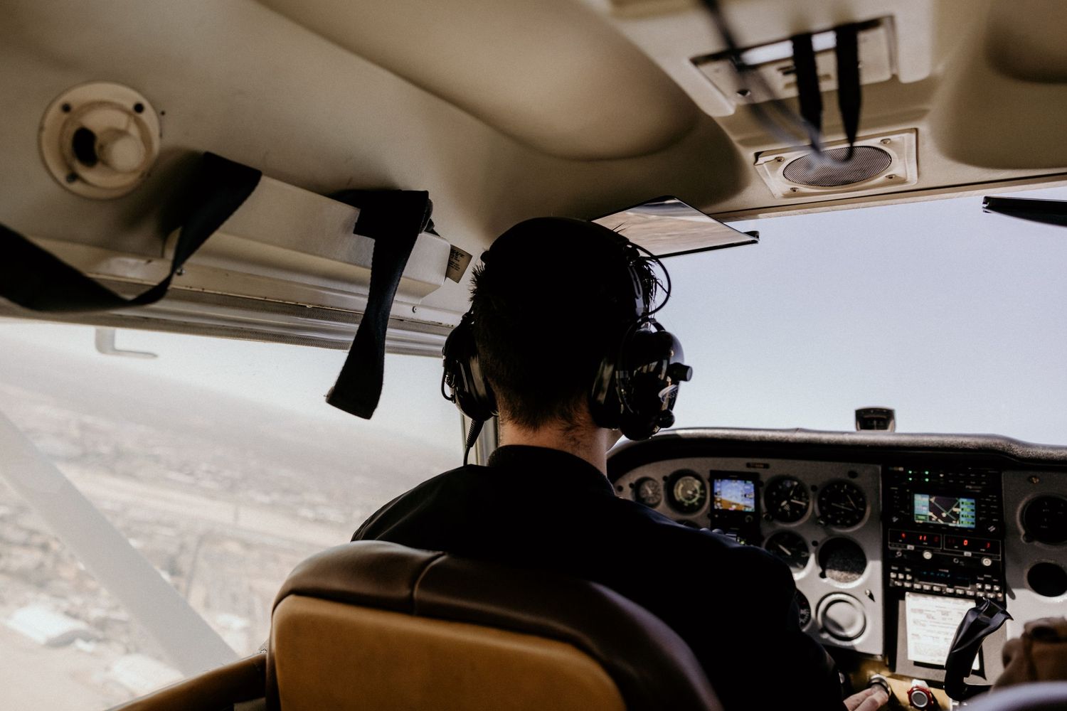 Rear view of pilot in headset flying small propeller plane over landscape.