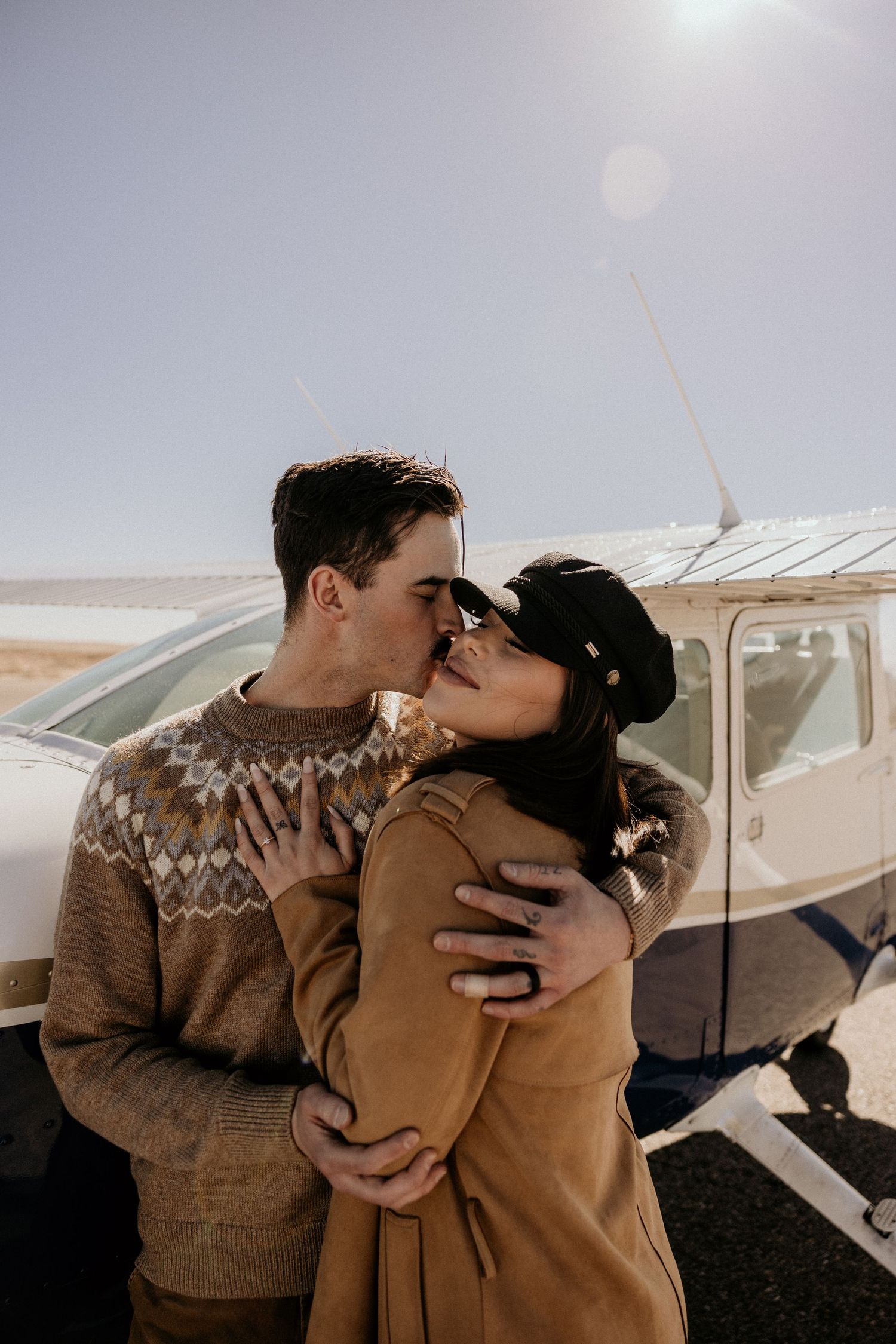 A couple shares a romantic kiss in front of a small private Cessna airplane outdoors.