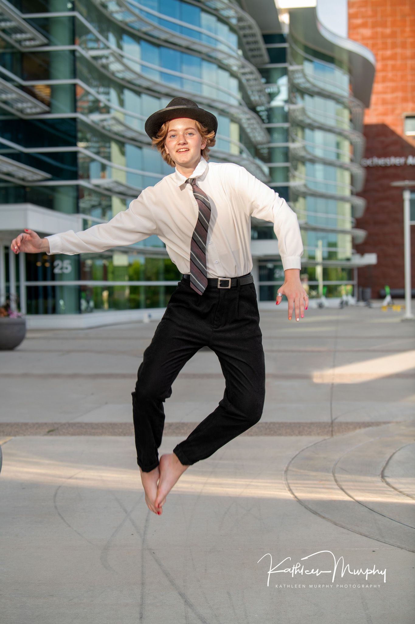 Energetic leap captured mid-air wearing business attire and hat outside modern building.