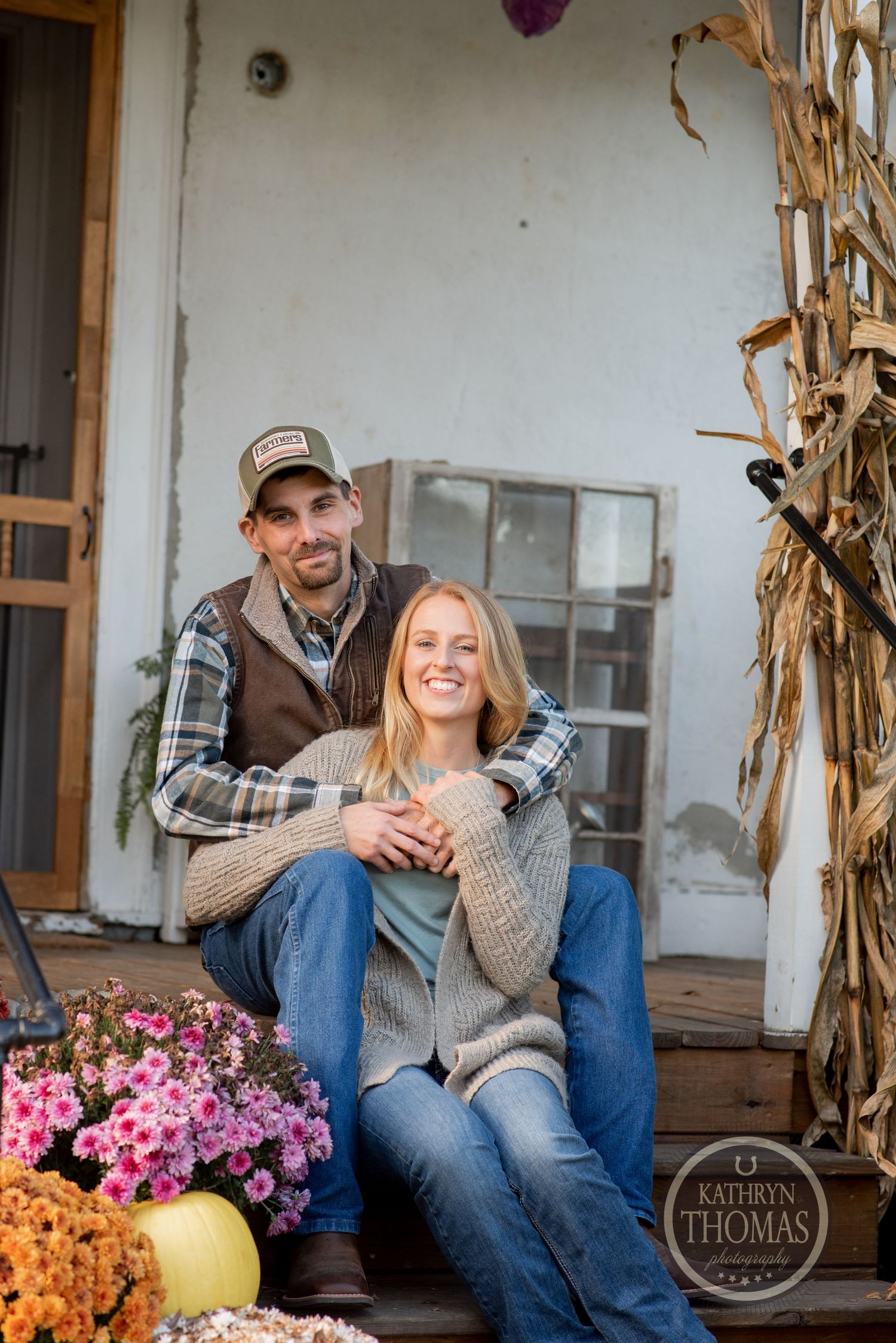 Couple sitting on rustic porch with autumn decorations including mums and corn stalks.