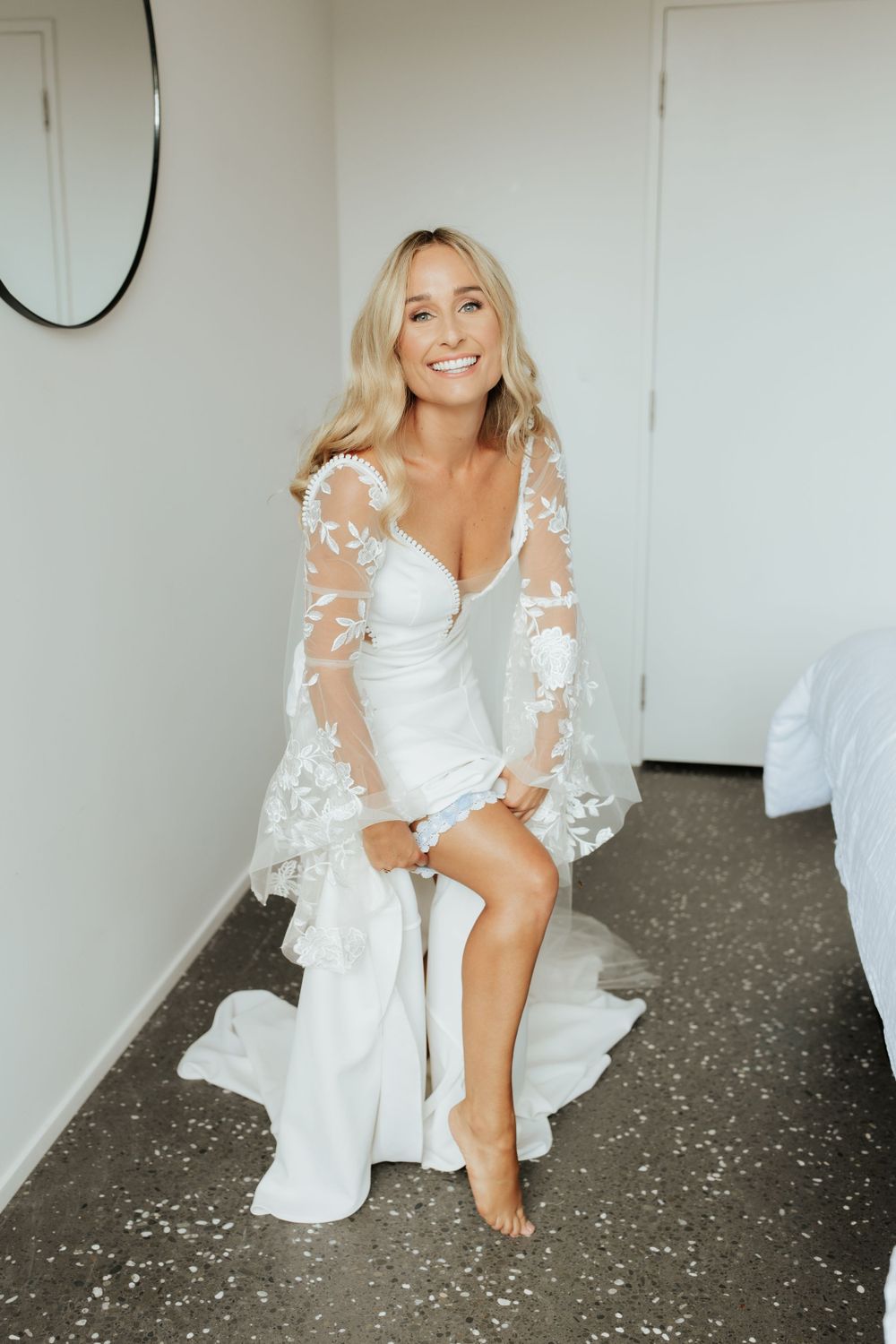 Bride in lace-sleeved wedding gown sitting elegantly in white room.