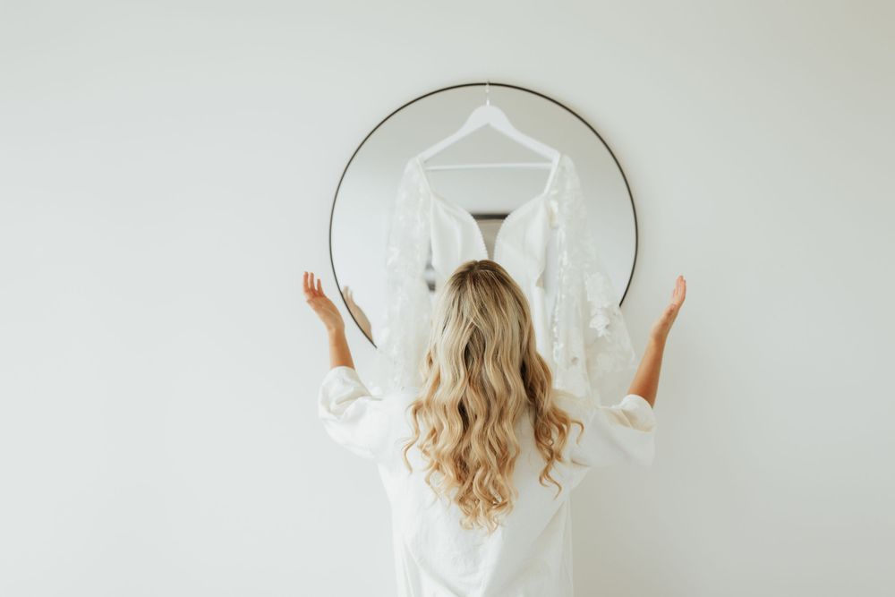 Wedding dress displayed on hanger reflected in round mirror against white wall.