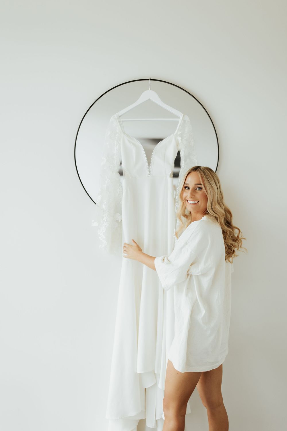 Wedding dress hanging on circular mirror while figure in white robe stands beside it.