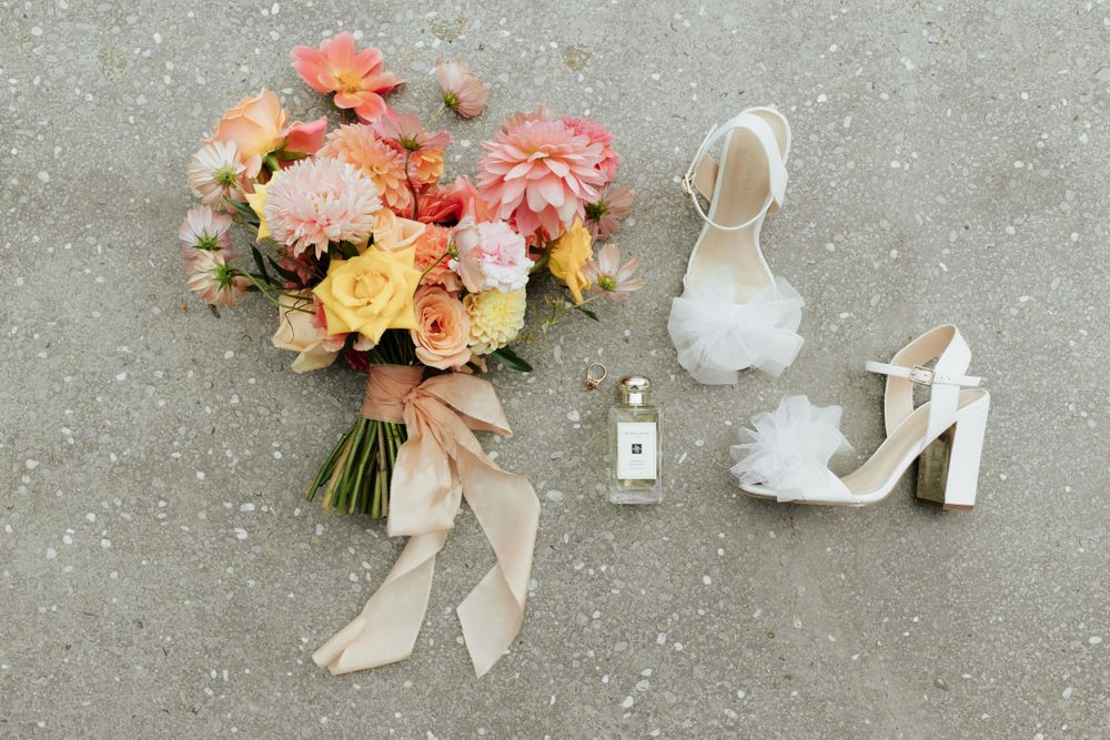 Wedding details flatlay with peach bouquet, white heels, and perfume bottle on concrete.