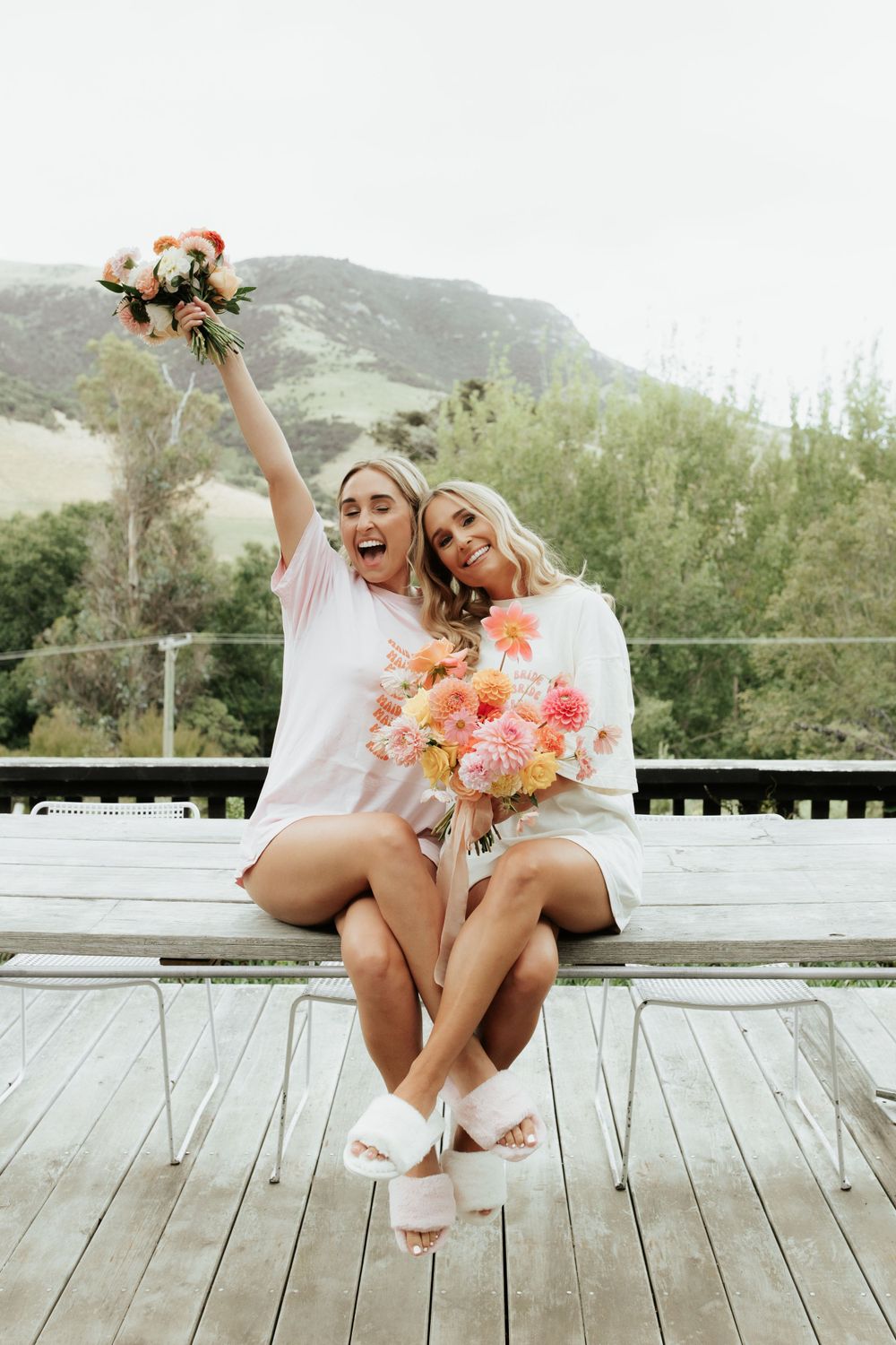 Two bridesmaids in white outfits sitting on wooden deck holding colorful bouquets with mountains in background.