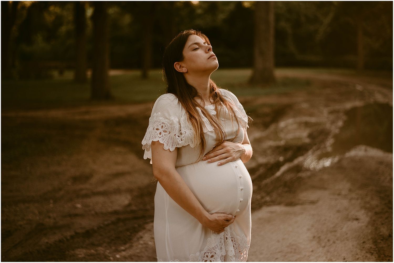 Ethereal maternity photo of woman in white lace dress cradling belly at golden hour on wooded path.