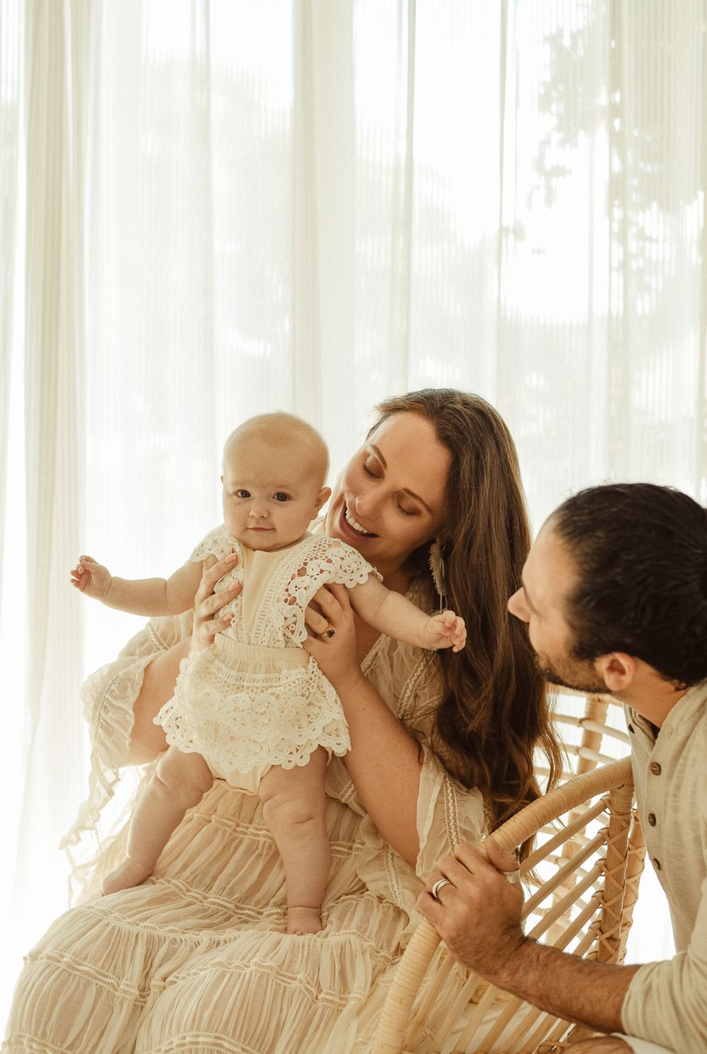 Family shares tender moment with baby wearing delicate white lace dress by sunlit window.