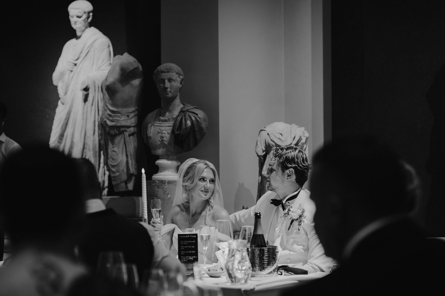 bride smiling at groom during father of the bride speech in the grand sculpture gallery at the Ashmolean Museum in oxford