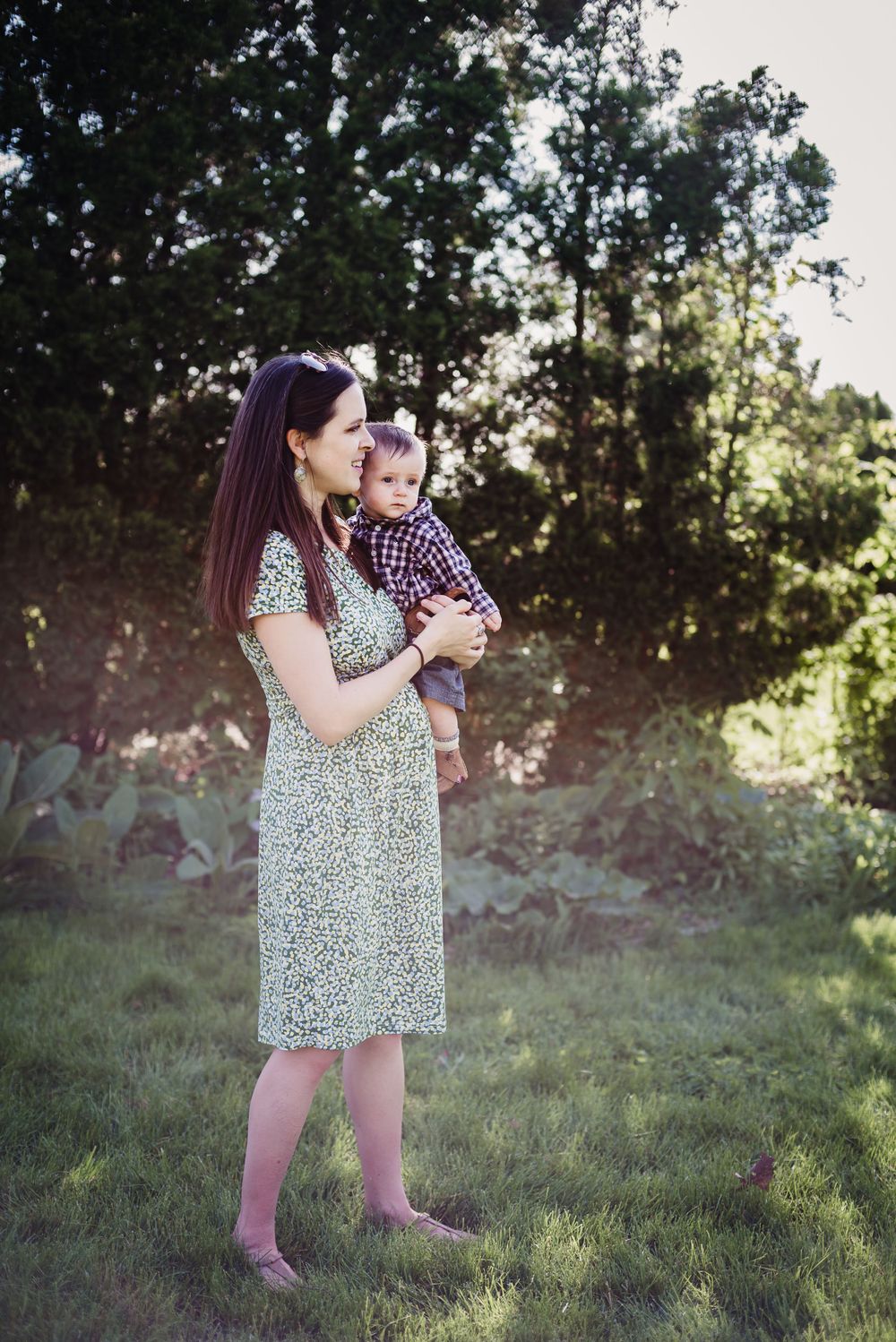 Colour image of a woman in a green dress holding her infant son as the sun shimmers in the trees behind her.