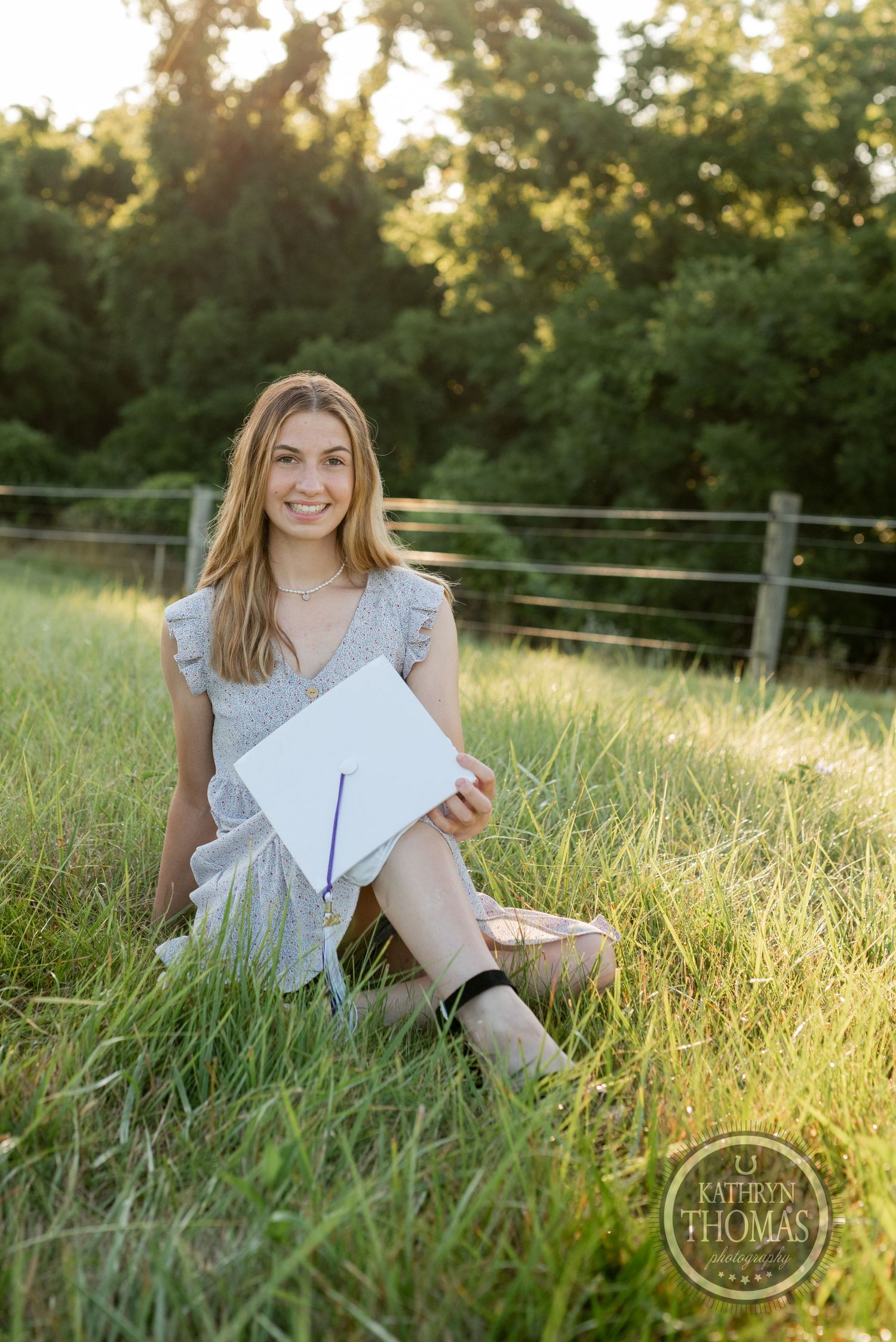 Graduate in grey dress holds diploma while sitting in sunlit field during golden hour.