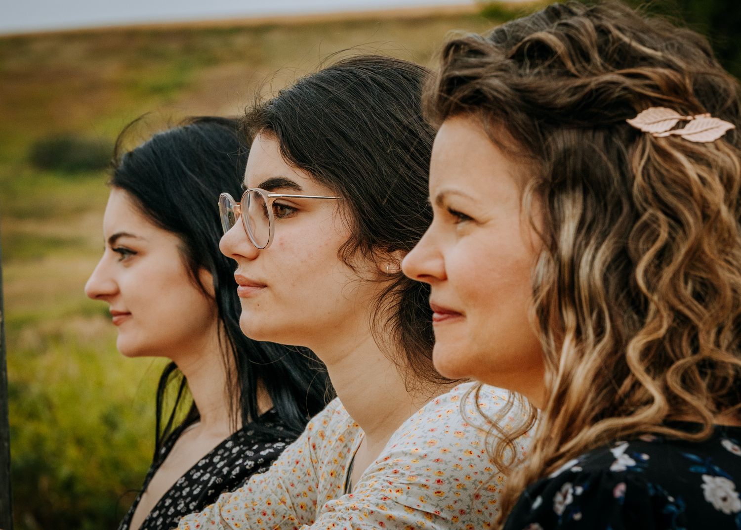 Three women with different hairstyles pose in profile against a natural outdoor backdrop.