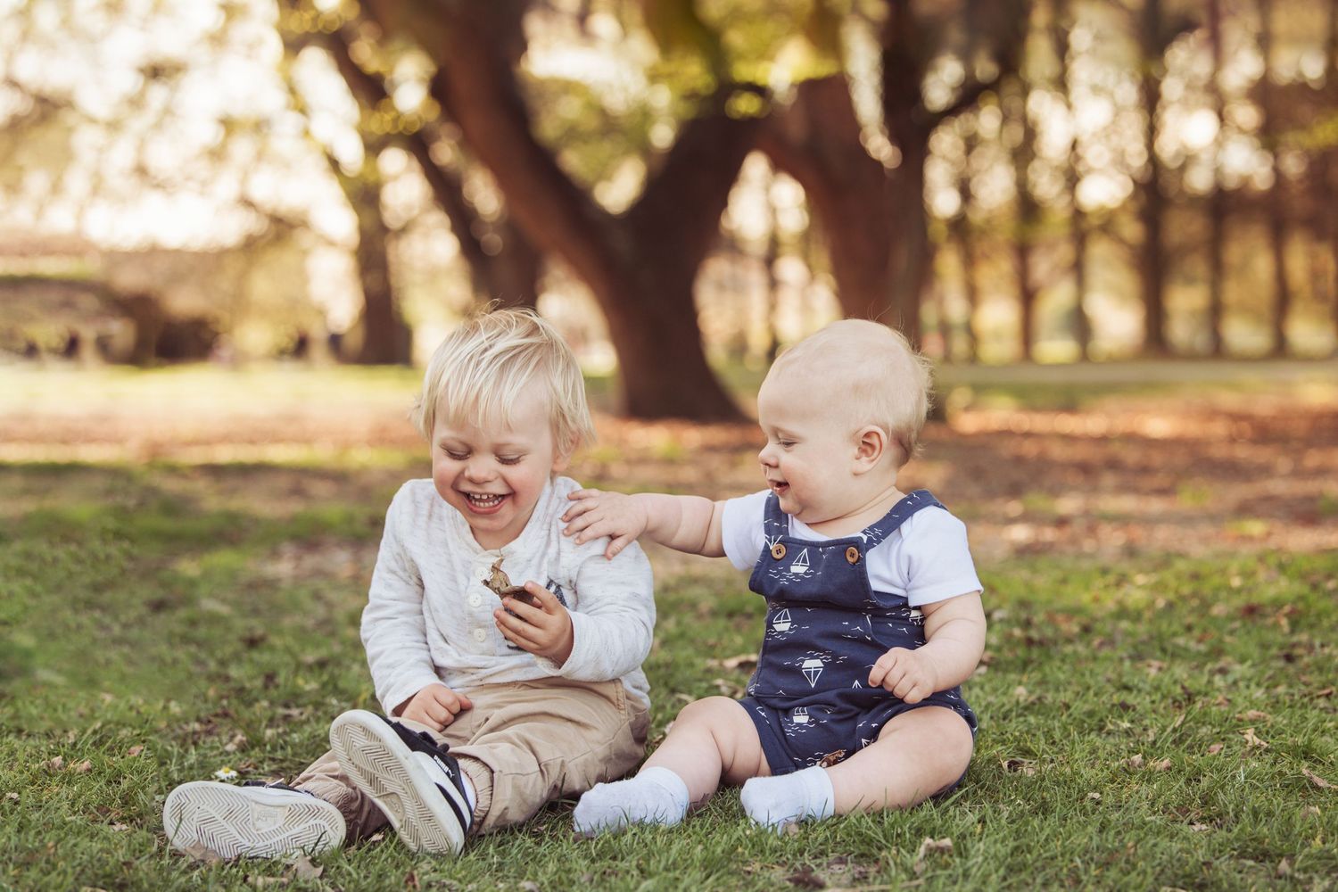 Two young children laugh together while sitting on grass during a sunny autumn day.