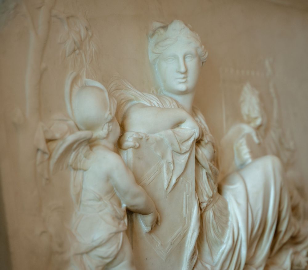 Classical marble relief sculptures depicting figures in flowing robes adorn a beige wall.