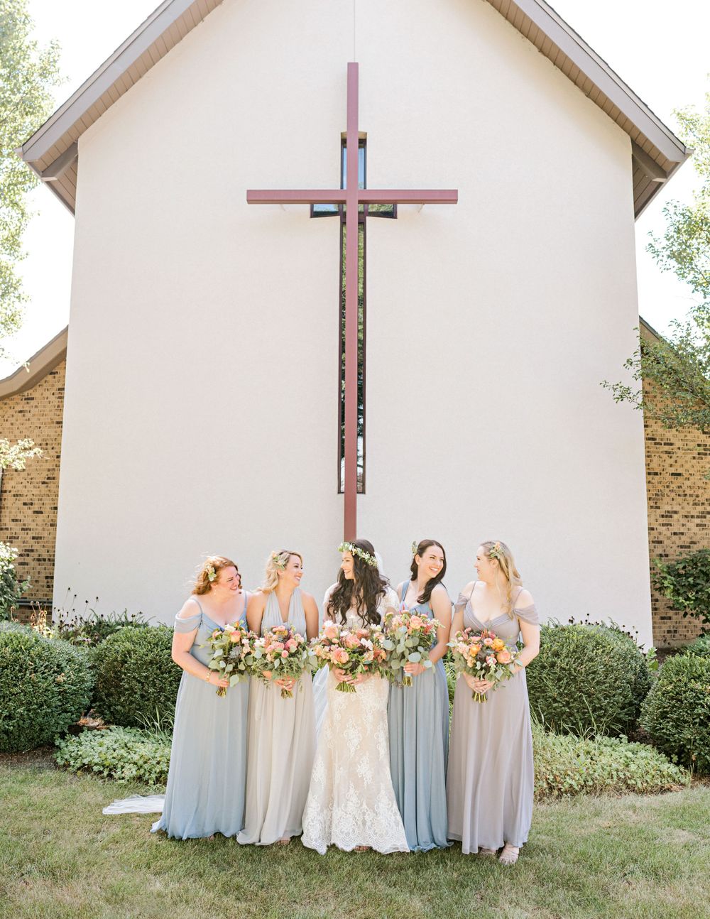 Bridal party in soft blue dresses posing in front of church with large cross.