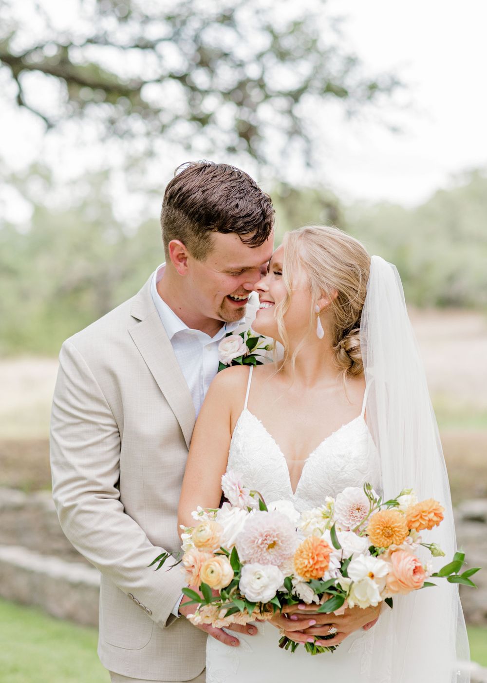 A tender wedding moment with couple in light attire sharing a close embrace while holding a peach and pink bouquet.