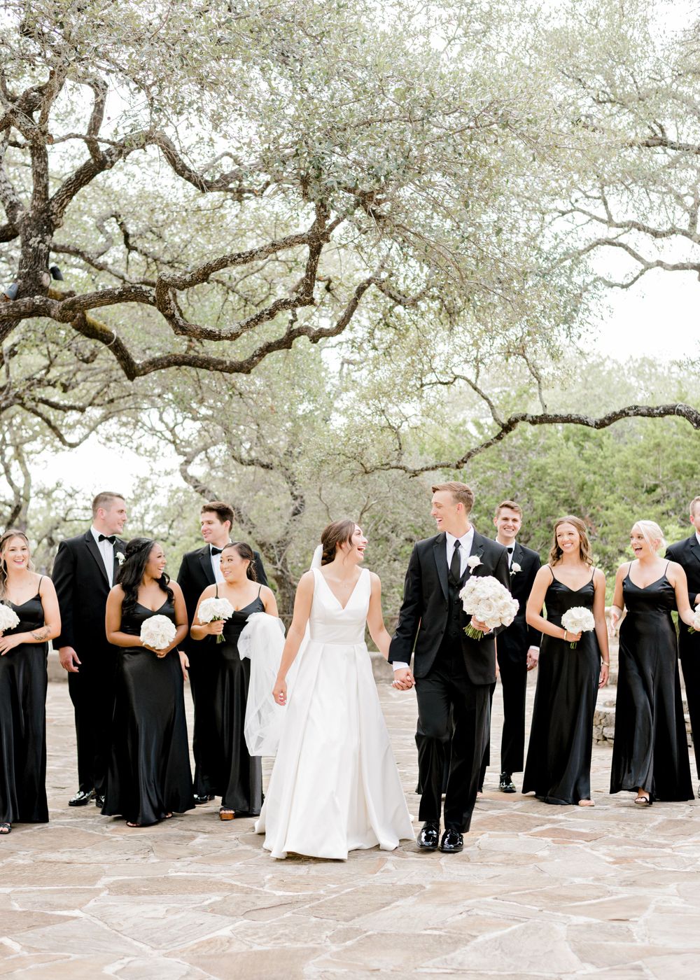 Wedding party in black and white attire poses under majestic oak trees.