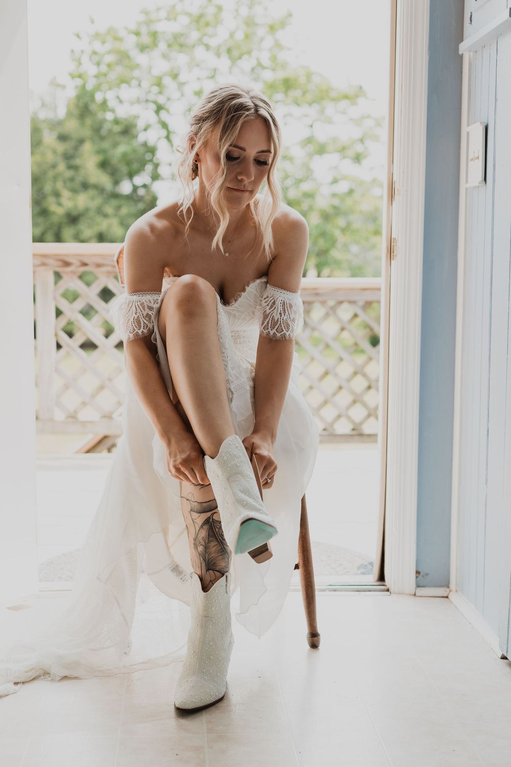 A bride in a lace dress putting on white cowboy boots while getting ready.