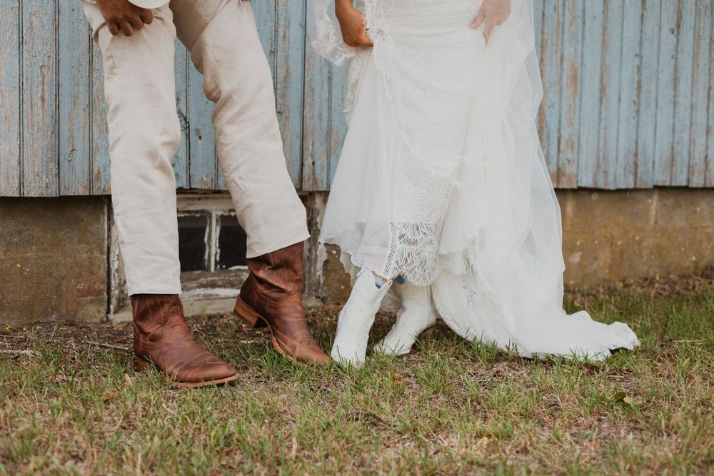 Close-up of brown leather boots and white wedding dress hem against weathered barn backdrop.