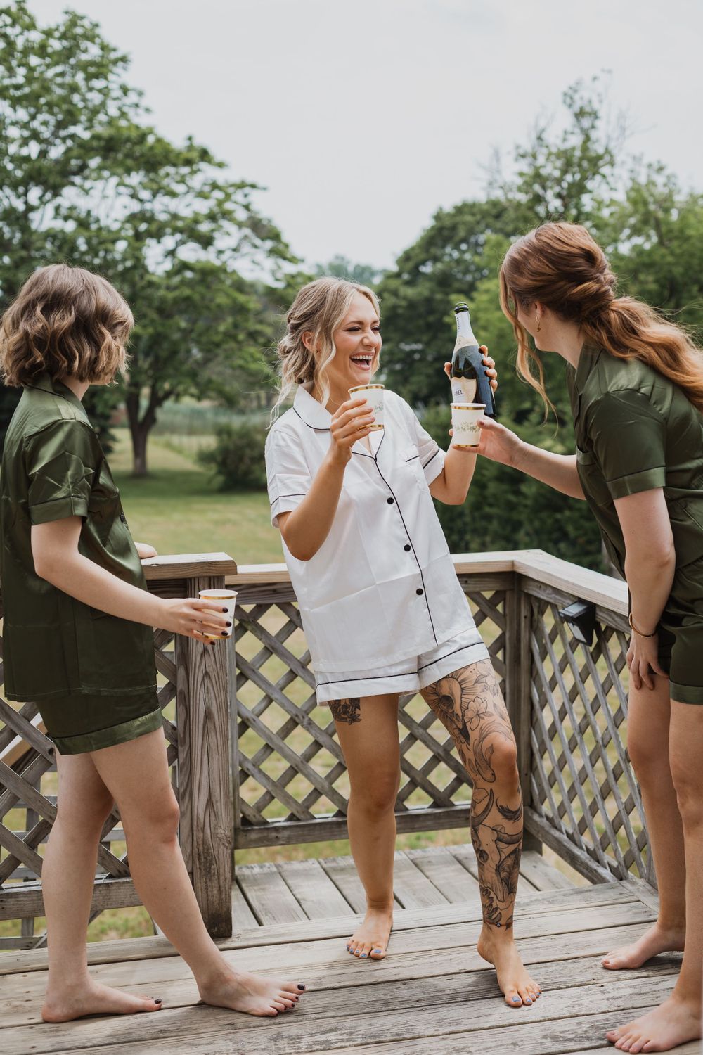 Three people in casual wear sharing drinks and laughs on wooden deck.