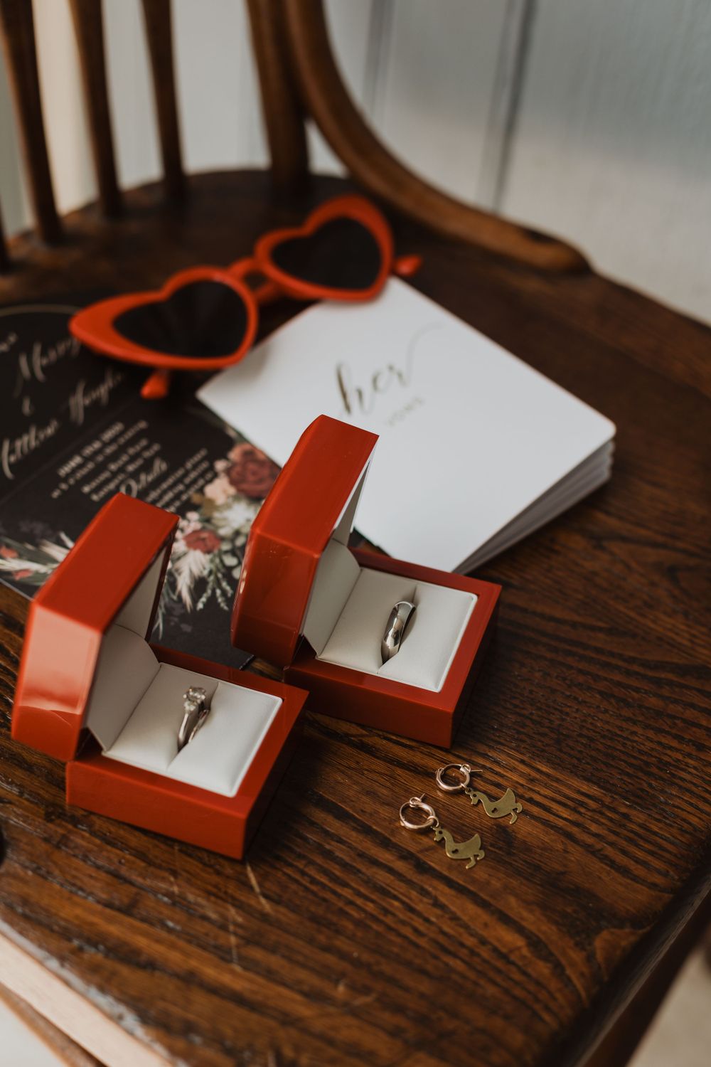 Wedding rings displayed in red boxes with heart-shaped sunglasses on wooden surface.