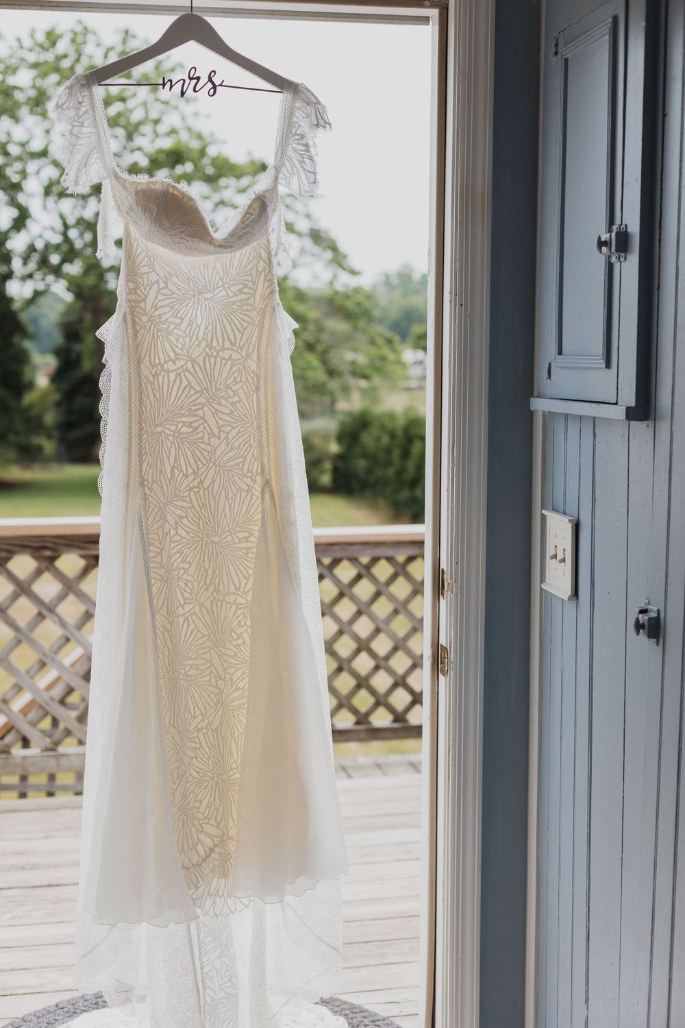 Elegant white lace wedding dress hanging by window with lattice deck visible outside.