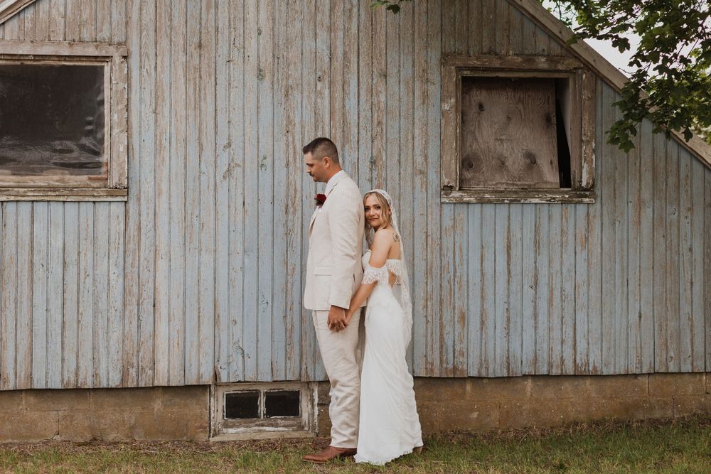 A couple poses in wedding attire against a rustic weathered barn backdrop.
