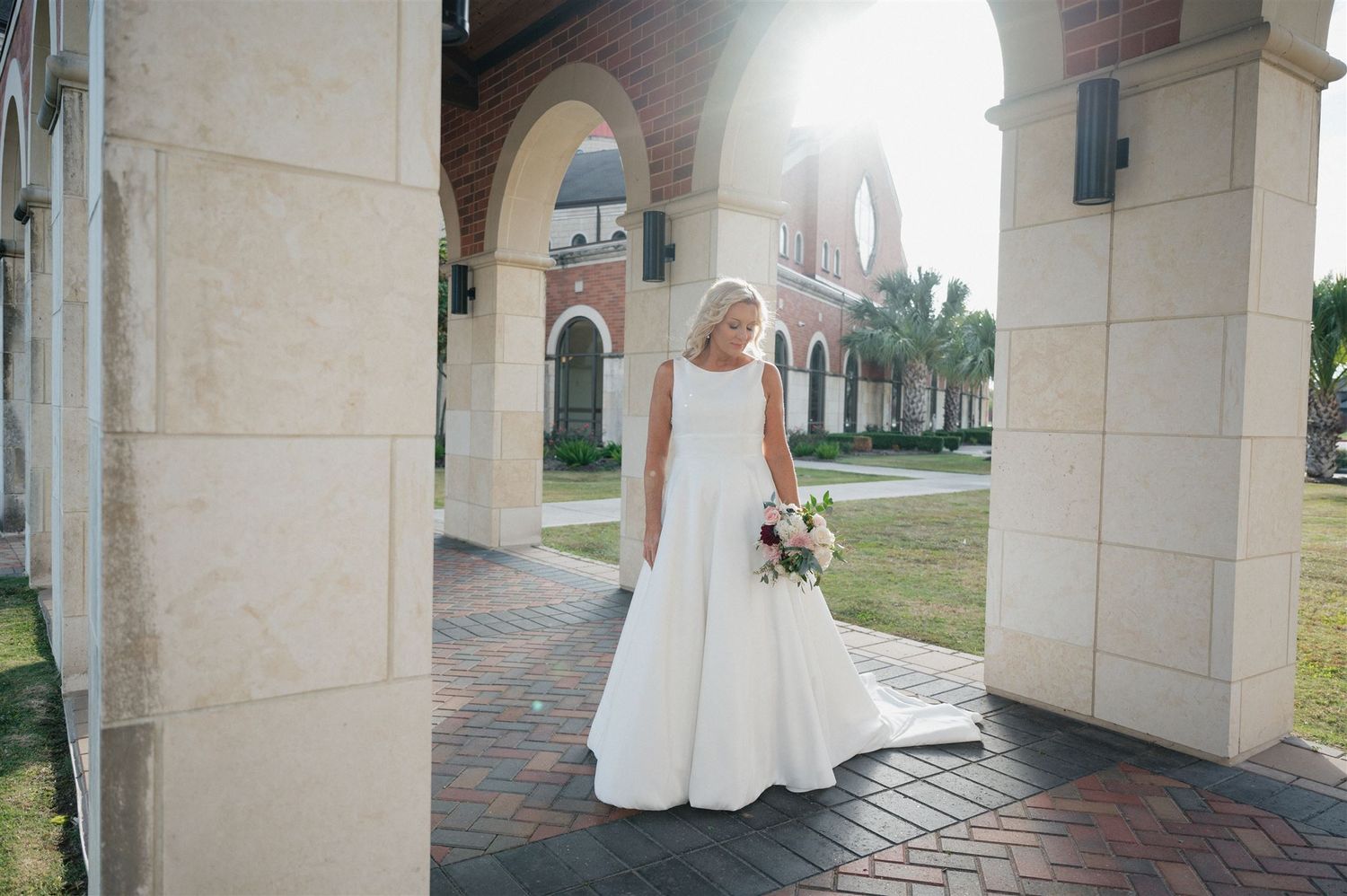 A bride in a white wedding gown stands under an arched colonnade holding a bouquet.