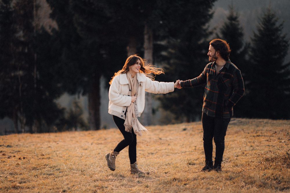 A couple dancing and holding hands on a grassy hillside surrounded by evergreen trees.