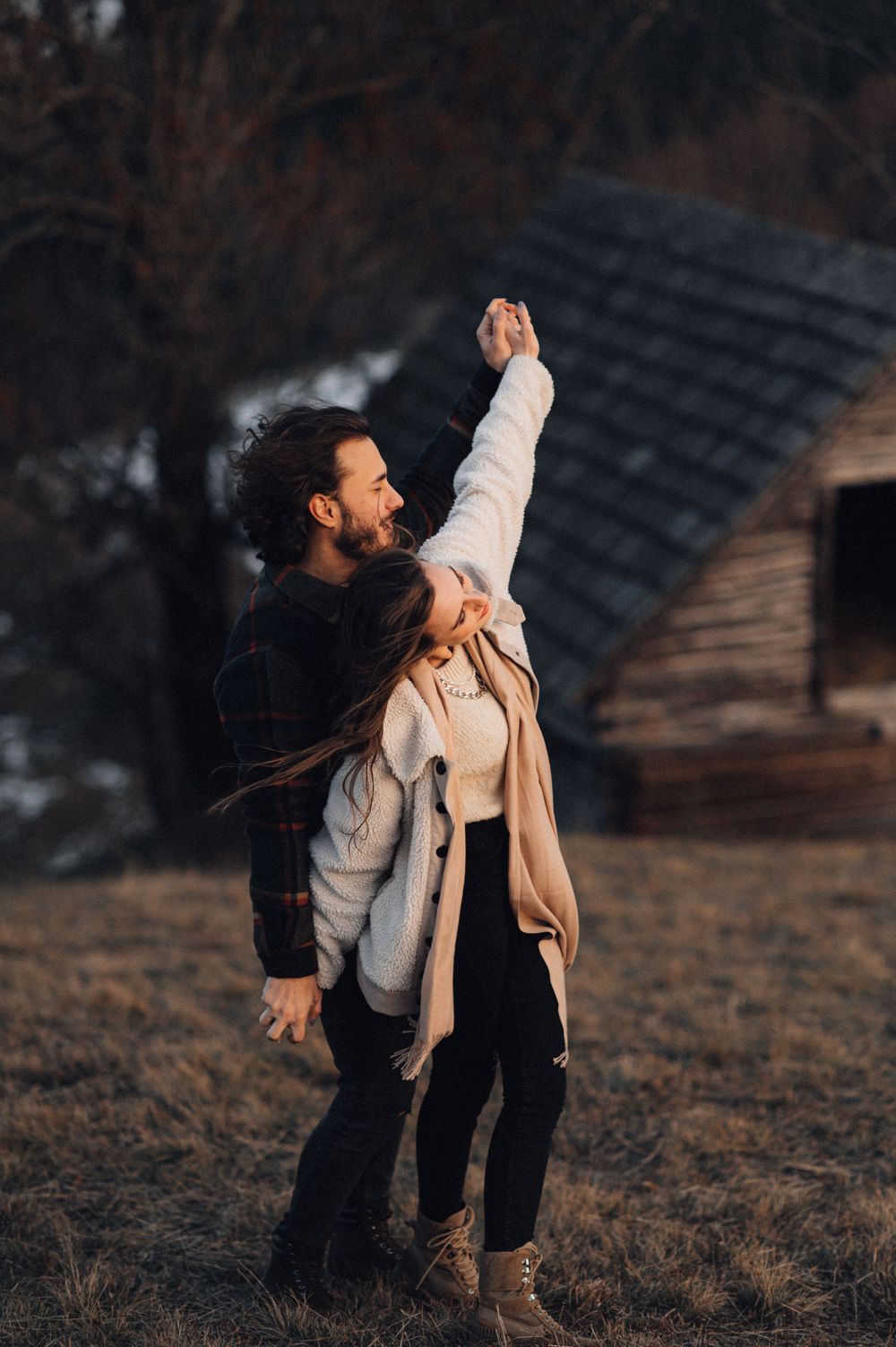 Couple dancing outdoors with arms raised near a rustic cabin at sunset.