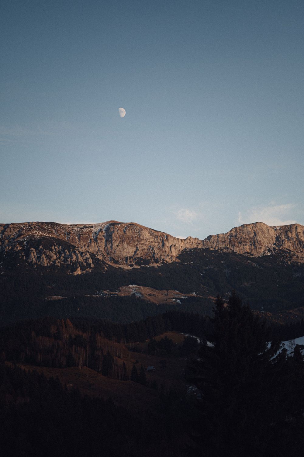 A scenic mountain landscape with a waxing moon visible in the clear blue sky.