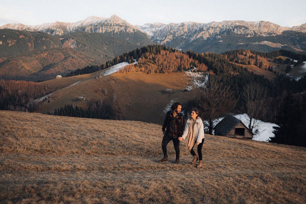 A couple stands together on a scenic mountain overlook with snowy peaks in the distance.