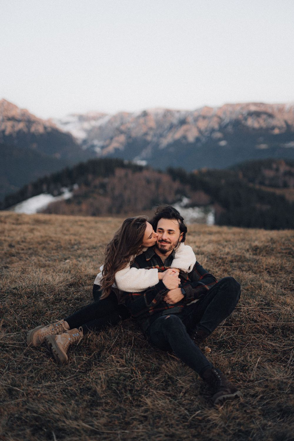A couple sits together on a grassy hill with snow-capped mountains in the distance.