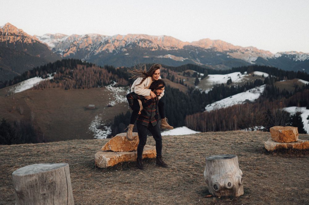 A couple embraces on a hilltop with snow-capped mountains and forests in the background.