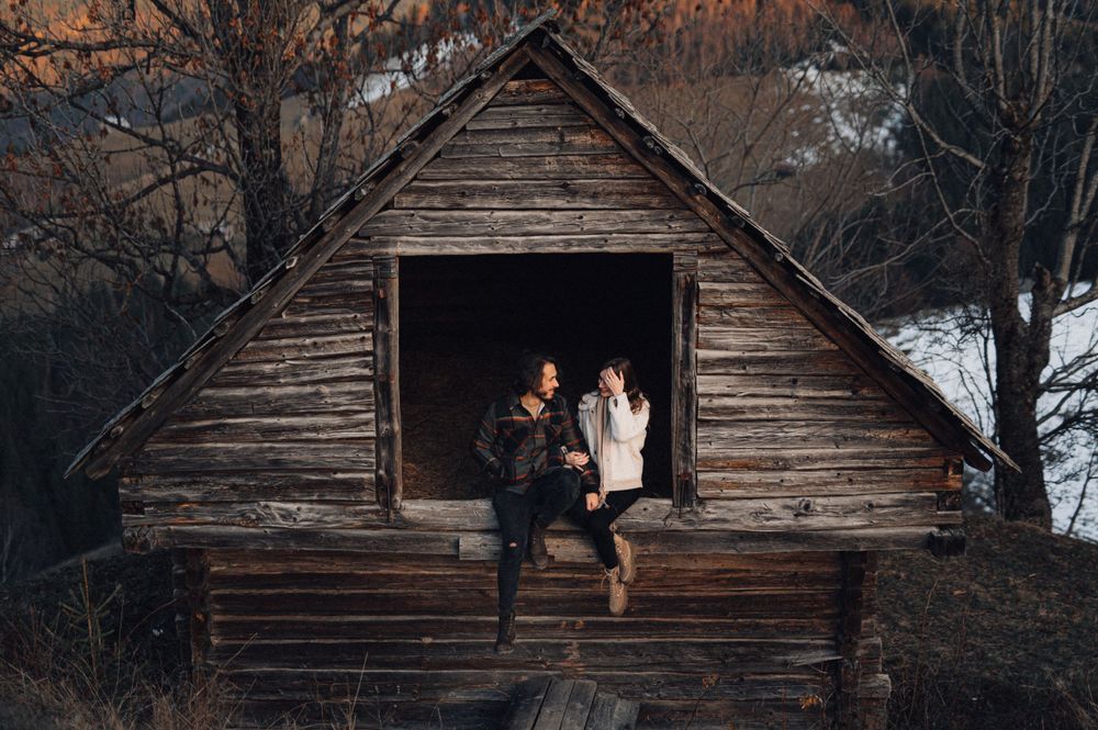 Couple sitting in doorway of rustic wooden cabin surrounded by winter landscape.