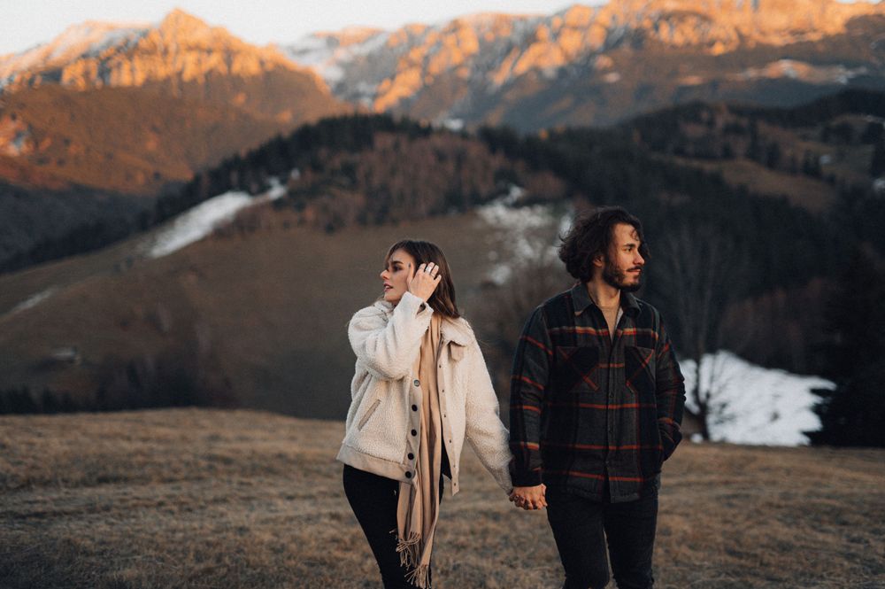Couple walking along mountain ridge with snow-capped peaks in background.