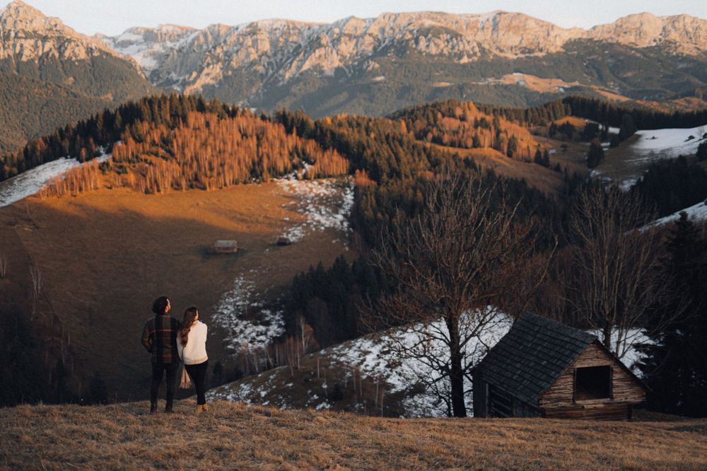 Two people stand together on a hill overlooking snow-capped mountains and autumn forests.