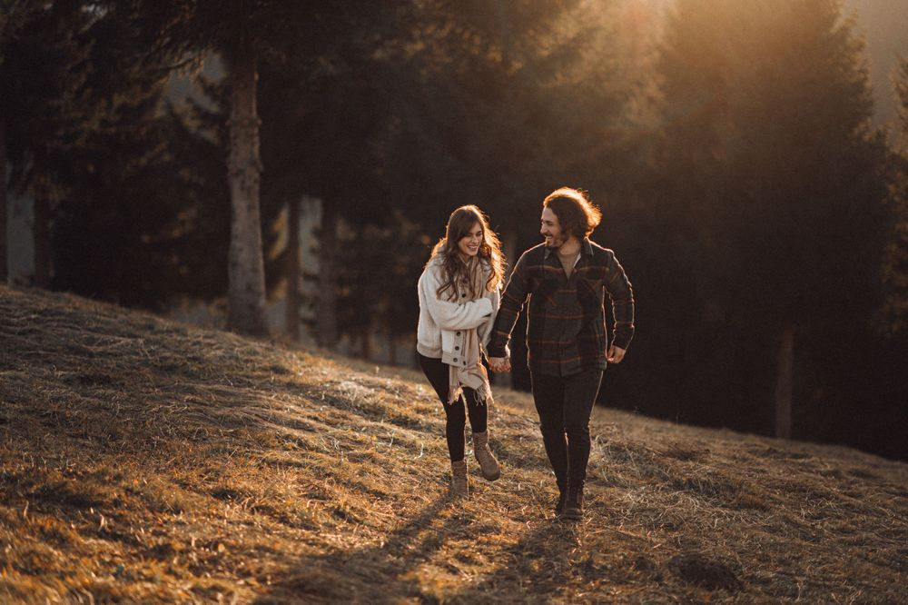 A couple walking together on a scenic hillside during golden hour with sunlight streaming through evergreen trees.